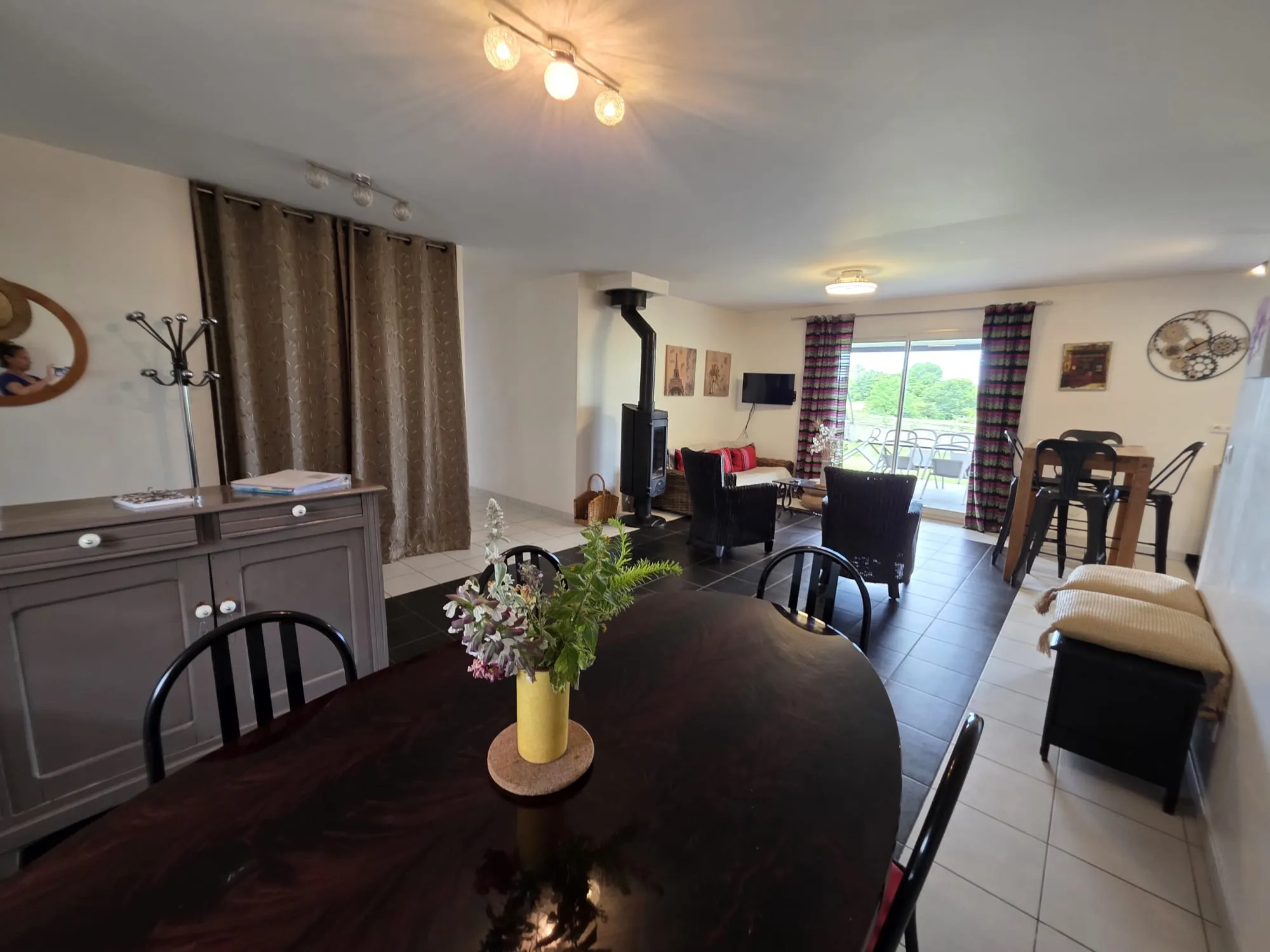 Vue d’ensemble de la pièce de vie : table à manger, poêle à bois et baies vitrées sur la terrasse